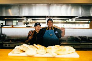 pexels-photo-6937411-6937411 Two chefs in a restaurant kitchen with baked goods, smiling and holding a rolling pin.